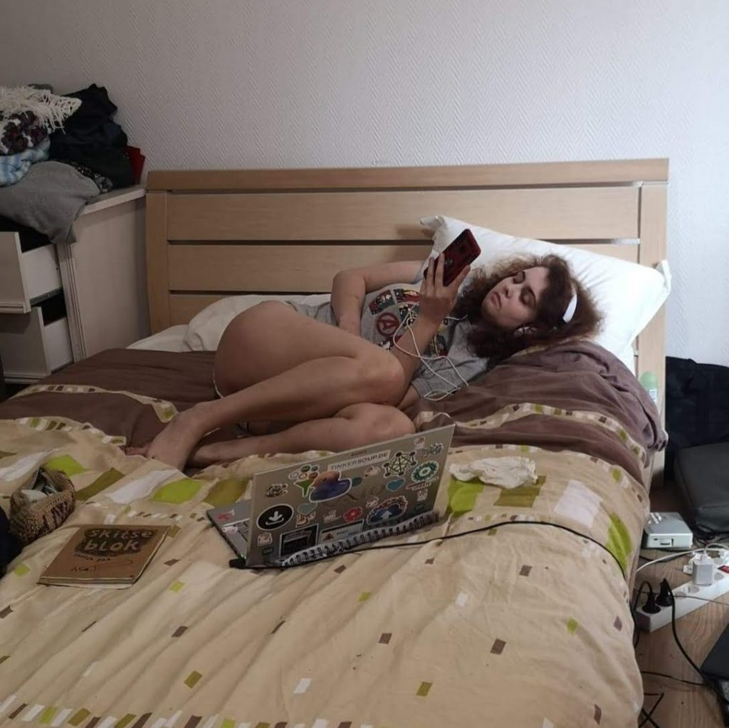 The author lies on a bed scrolling on her phone, headphones on, laptop and sketchbook in front of her